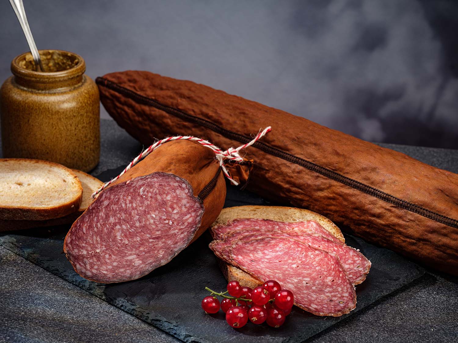 Frische Bauernsalami (geräuchert): Eine aufgeschnittene Bauernsalami liegt mit drei Brotscheiben und Senf, angerichtet auf einer schwarzen Schieferplatte