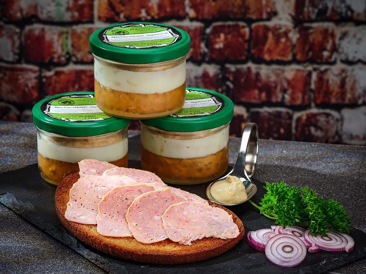 Mechterstädter Jagdwurst im Glas: Auf einer gerösteten Brotscheibe liegen mehrere Stücke Jagdwurst, angerichtet mit Petersilie, Zwiebel und Senf auf einer schwarzen Schieferplatte. 3 Gläser Jagdwurst stehen im Hintergrund