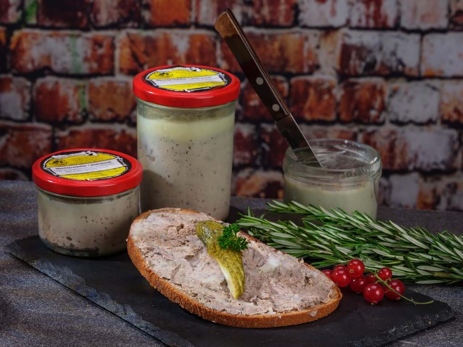 Mechterstädter Leberwurst im Glas: Eine Scheibe Brot mit Leberwurst, angerichtet auf einer schwarzen Schieferplatte, mit Gurke und Roten Johannisbeeren. Dahinter stehen drei Gläser Leberwurst