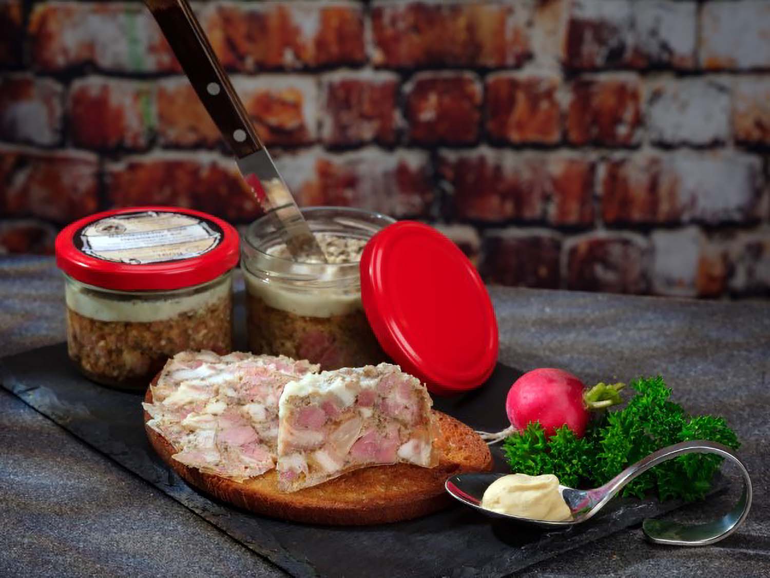 Mechterstädter Sülze im Glas: Auf einer gerösteten Brotscheibe liegen mehrere Stücke Sülze, angerichtet mit Petersilie und einem Radieschen auf einer schwarzen Schieferplatte. 2 Gläser Sülze stehen im Hintergrund