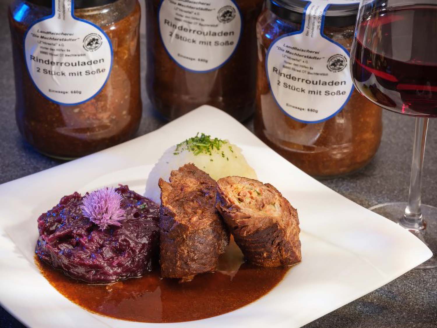 Rinderrouladen (2 Stück): Eine Portion Rinderrouladen, angerichtet auf einem Teller mit Kloß und Rotkohl. Dahinter stehen drei Gläser Rinderrouladen, sowie ein Glas Rotwein