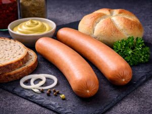 Ein Paar Bockwurst liegt, zusammen mit einem Brötchen, Senf und zwei Scheiben Brot, dekorativ angerichtet auf einer schwarzen Schieferplatte